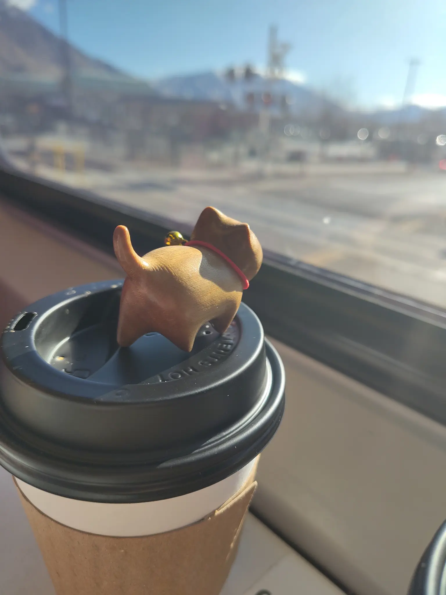 a tiny wooden carved cat with a bell on its neck, sitting on a coffee cup and looking out the window of a train.